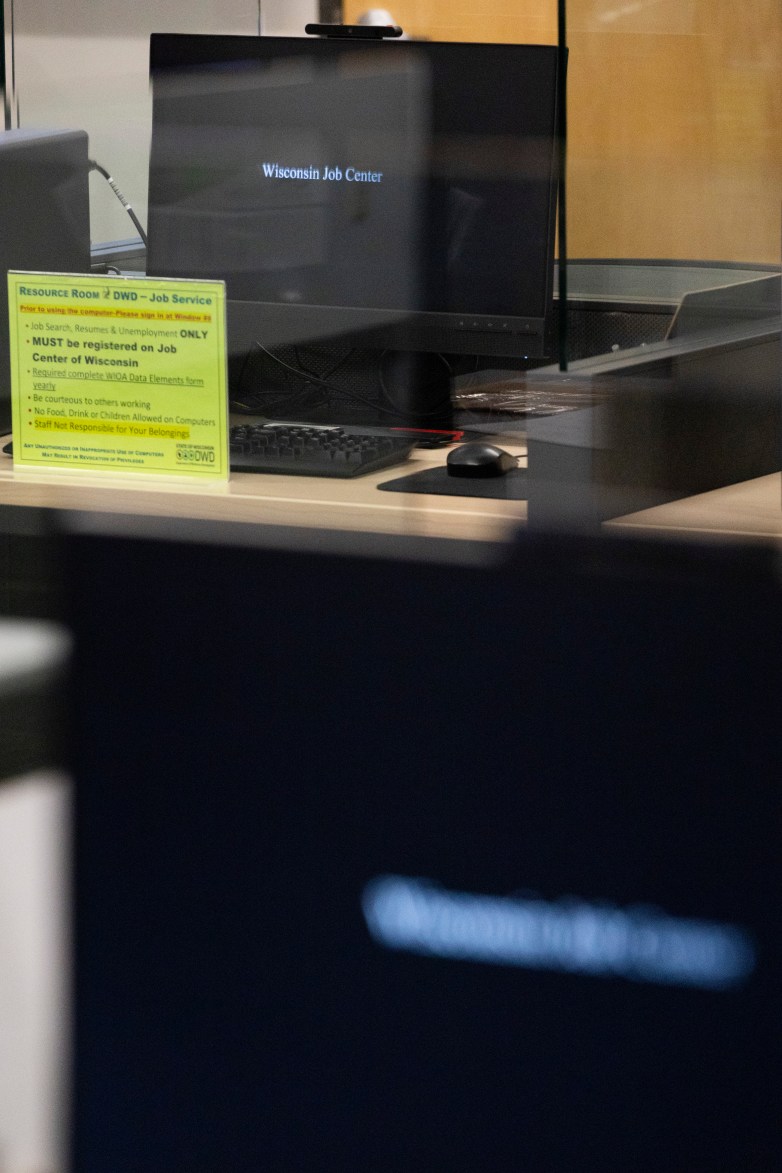 A computer workstation sits on a desk, with a monitor displaying “Wisconsin Job Center” and a yellow sign listing job service and computer use rules.