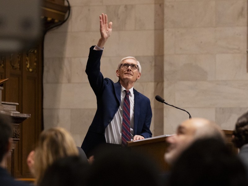 A person stands and raises a hand at a podium with a microphone in a marble-walled room, with other people sitting in the foreground.