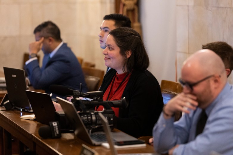 People sit at a wooden desk with laptops and a video camera on the desk.