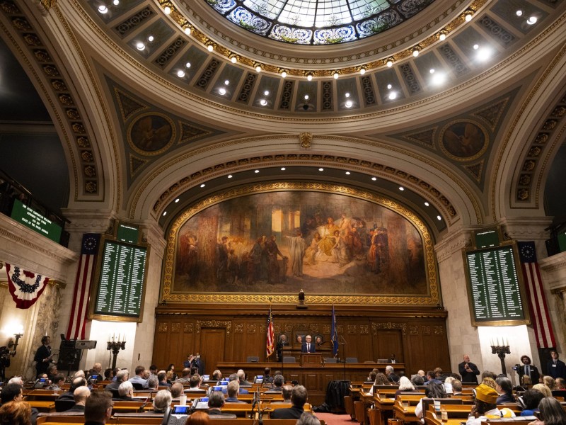 A wide view of a legislative chamber shows people seated at desks facing a person at a podium beneath a large mural, with flags behind the podium and electronic voting boards on the walls.