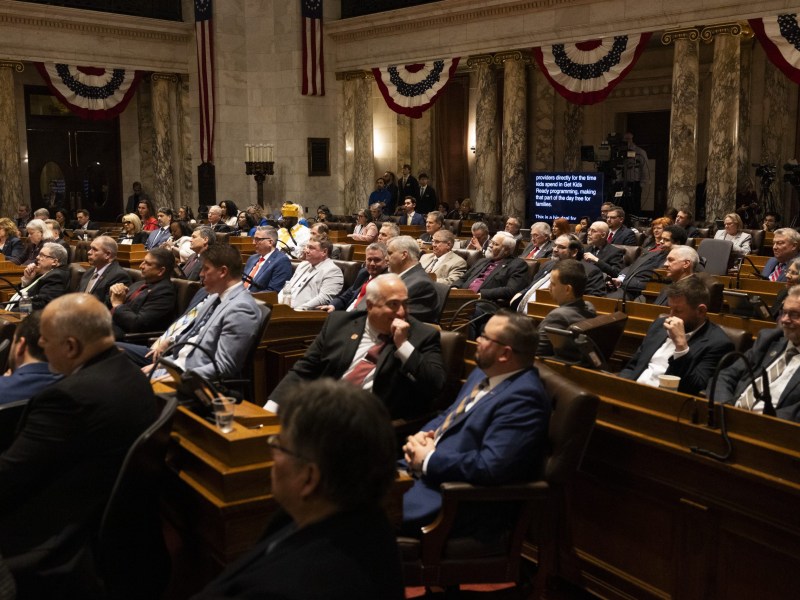 Several people sit at wooden desks in a marble-columned room decorated with red, white and blue bunting.