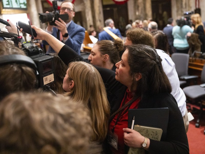 People gather closely around cameras and microphones in a room while a person holds a notebook, pen and smartphone in the foreground.