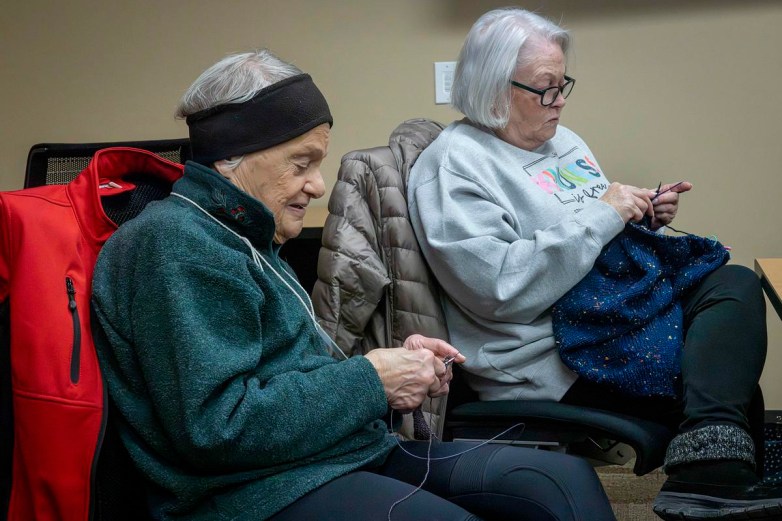 Two people sit in chairs knitting with needles and yarn, with coats draped over the backs of chairs inside a room.