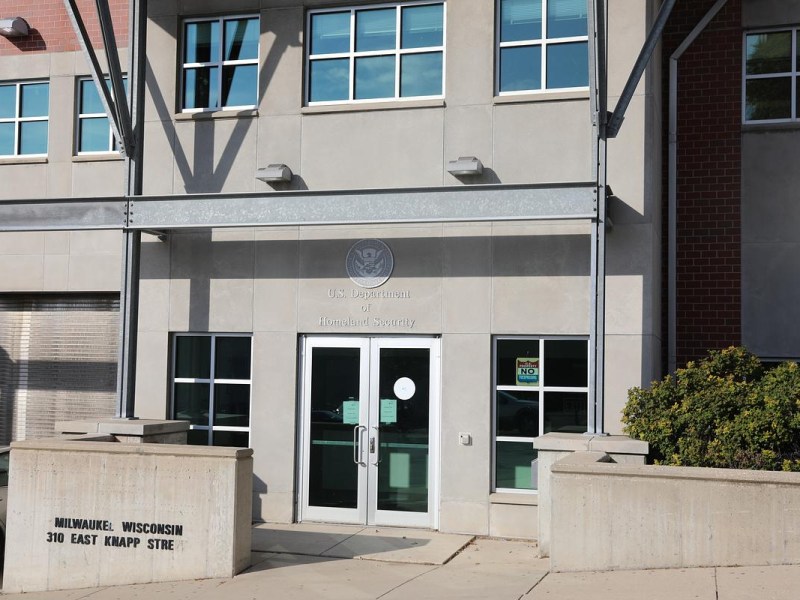 Entrance of a gray concrete building with "U.S. Department of Homeland Security" above glass doors and "Milwaukee, Wisconsin 310 East Knapp St" on a concrete sign in front.