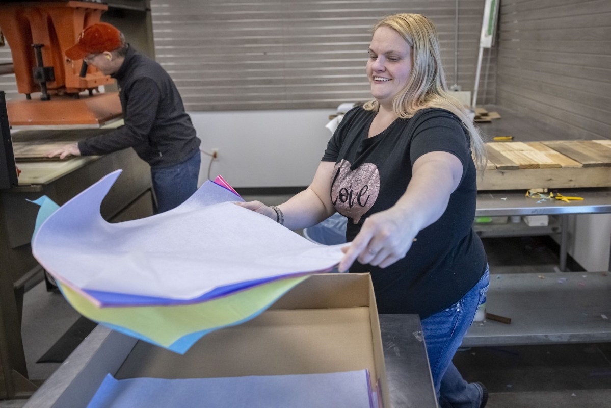 A person places sheets of colorful material into a cardboard box at a worktable while another person stands at another worktable in the background.