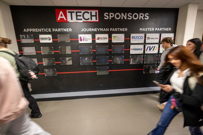 People walk past a wall display reading “ATECH Sponsors” with sections labeled “Apprentice Partner,” “Journeyman Partner” and “Master Partner,” showing multiple company logos and empty plaques.
