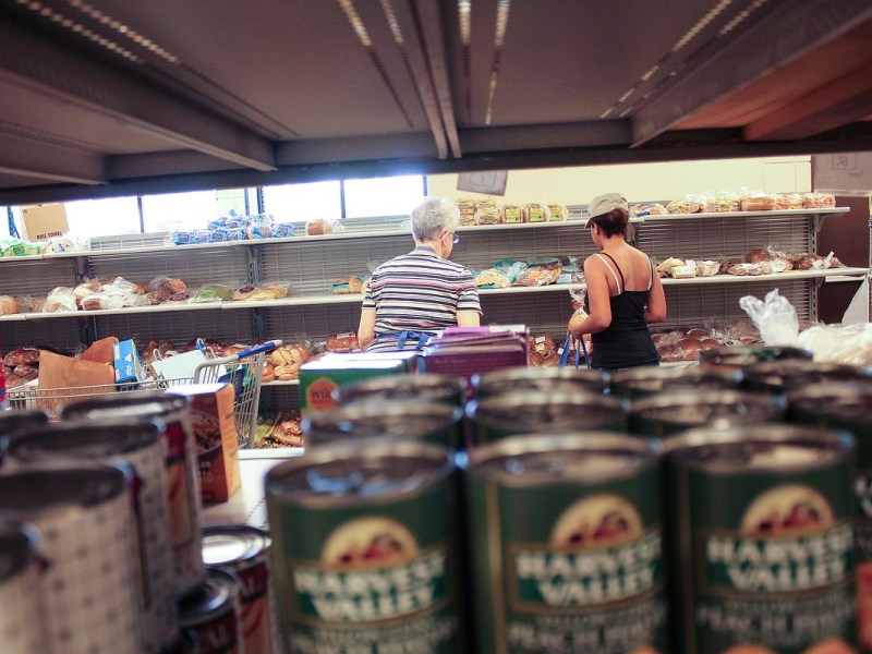 Two people stand near mostly empty bread shelves with a shopping cart visible, seen from behind rows of canned goods in the foreground.