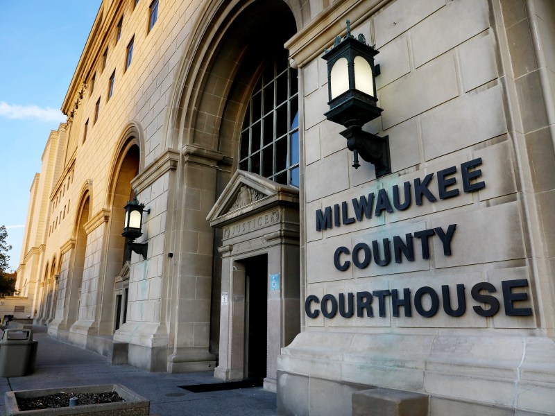 The exterior of a building shows large arched windows, stone walls and a sign reading "MILWAUKEE COUNTY COURTHOUSE" next to an entrance with the word "JUSTICE" above a door.