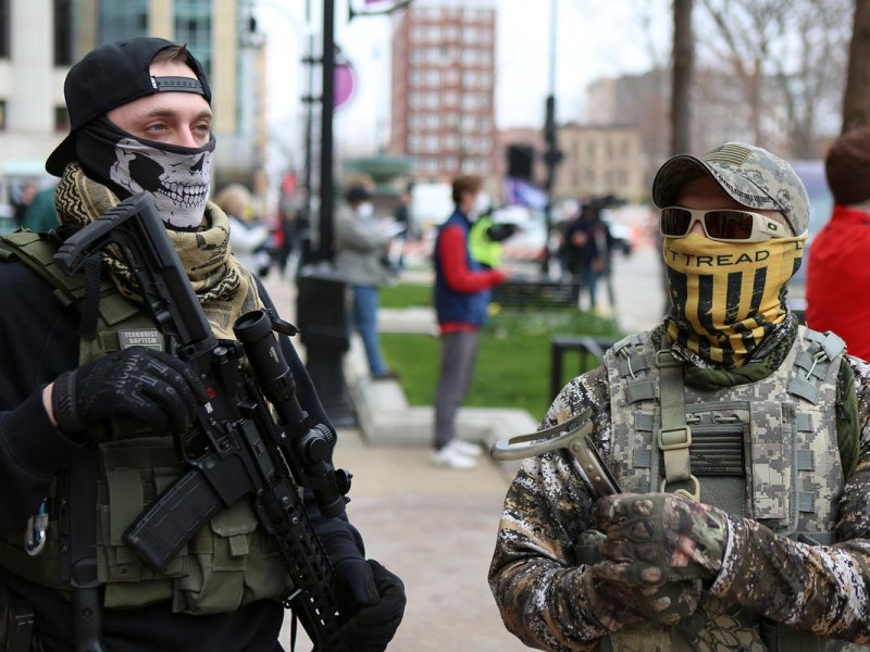 Two people wearing face coverings and tactical-style clothing stand outdoors, one holding a rifle, with other people blurred in the background.
