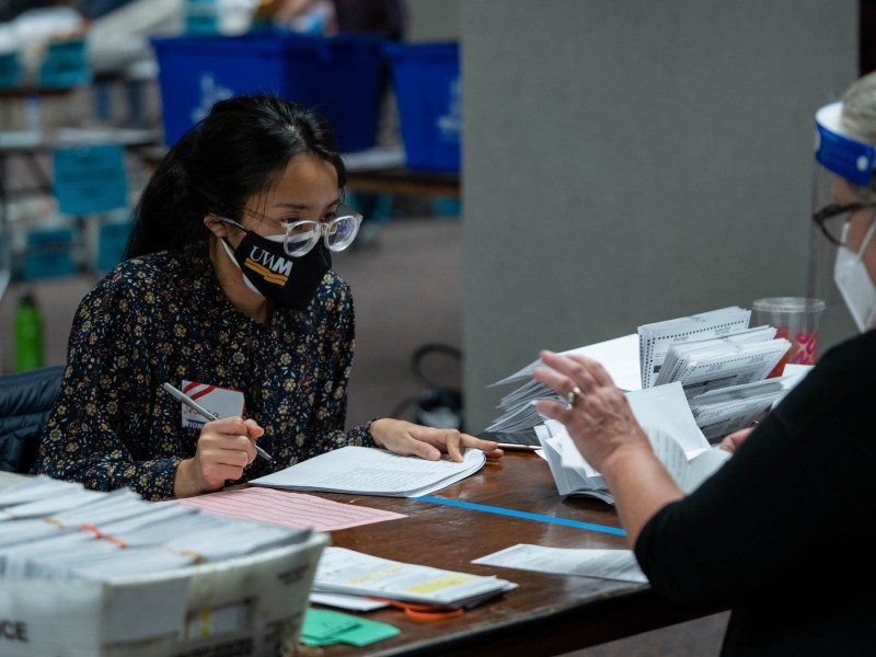 People wearing masks and glasses, one of them wearing a face shield, look at and hold pieces of paper at a table, with envelopes, forms and a tray labeled “United States Postal Service” visible on the table.