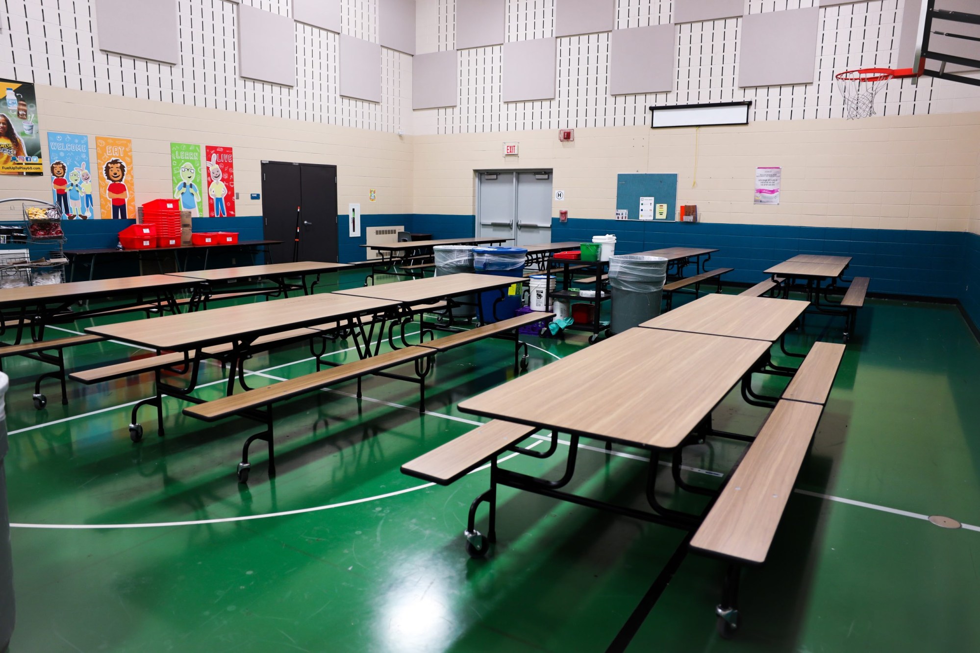 An empty room with long tables, attached benches and a green floor, with colorful posters and a basketball hoop on the walls.