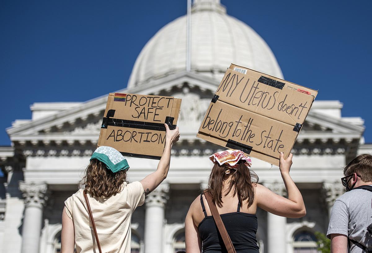 People hold cardboard signs reading "PROTECT SAFE ABORTION" and "MY Uterus doesn’t belong to the state" outside a white domed building under a clear blue sky.