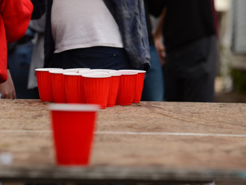 A group of red plastic cups arranged in a triangle sits on a wooden table, with people standing with faces unseen in the background.