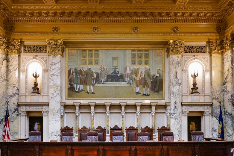 A row of wooden chairs and microphones sits beneath marble walls and a large framed painting of people gathered in a historical interior.