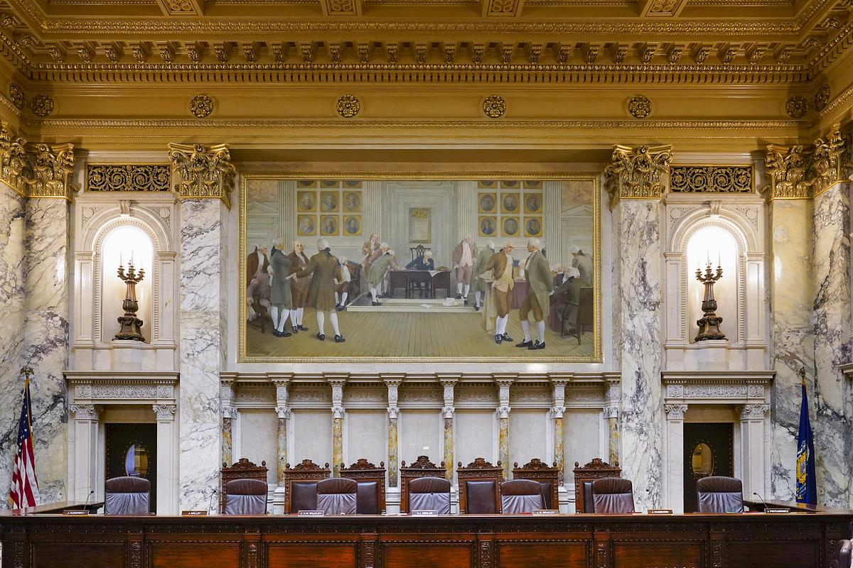 A row of wooden chairs and microphones sits beneath marble walls and a large framed painting of people gathered in a historical interior.