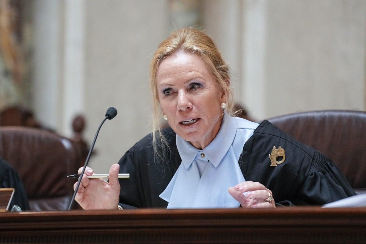 A person in a black and blue robe sits in a chair and holds a pen near a microphone.
