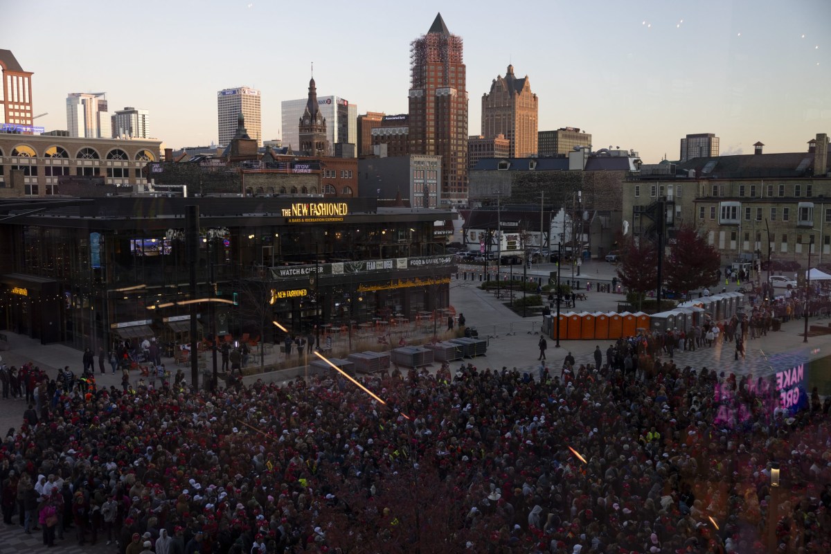 A large crowd gathers in a downtown plaza near a building with a sign reading "THE NEW FASHIONED," with high-rise buildings and a city skyline in the background.