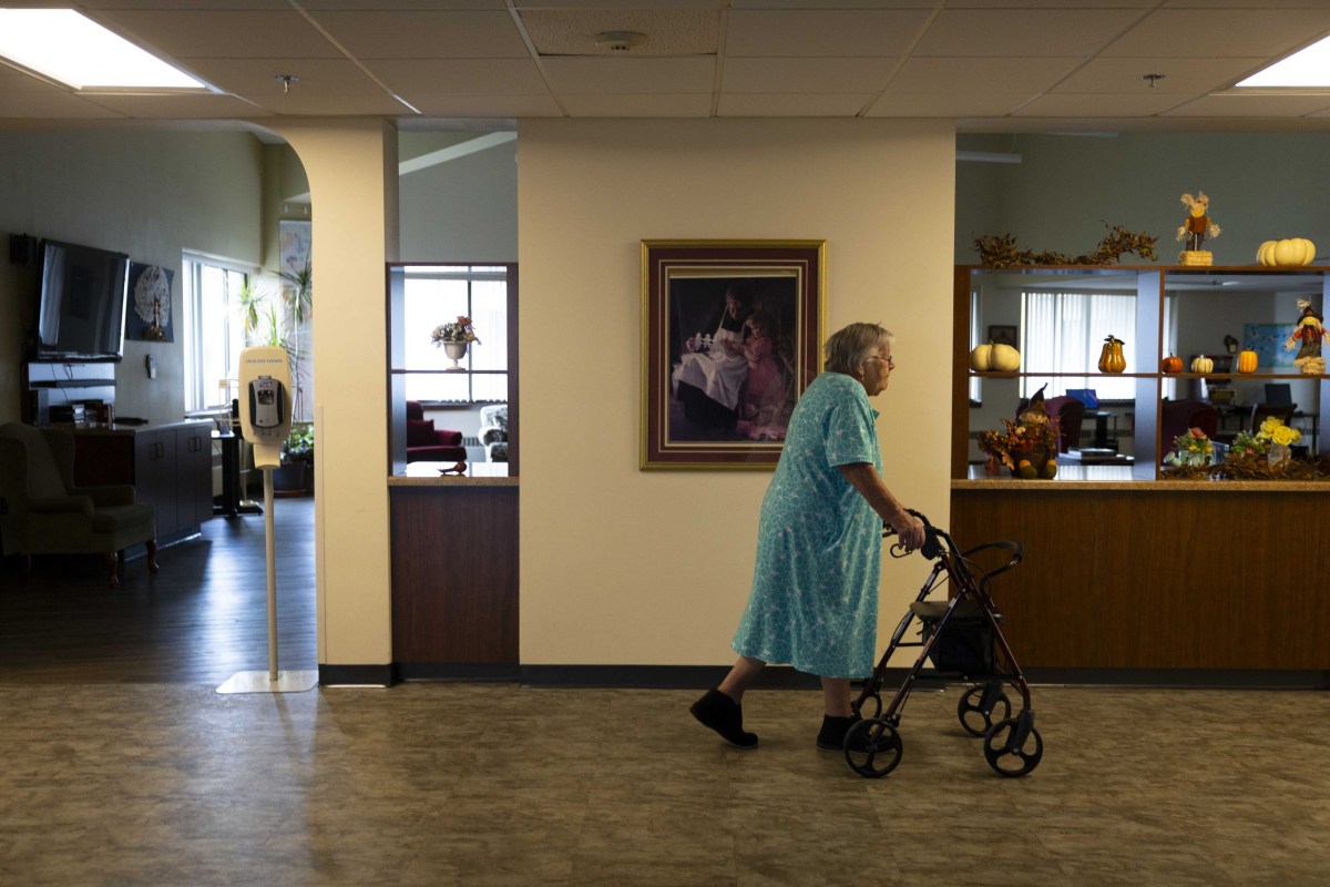 A person in a blue patterned dress walks with a wheeled walker in a hallway, with pumpkins and autumn decorations on shelves and a framed painting on a wall and a room visible behind the wall.