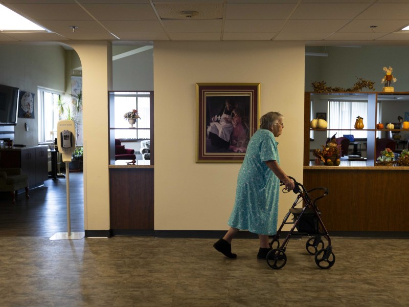 A person in a blue patterned dress walks with a wheeled walker in a hallway, with pumpkins and autumn decorations on shelves and a framed painting on a wall and a room visible behind the wall.