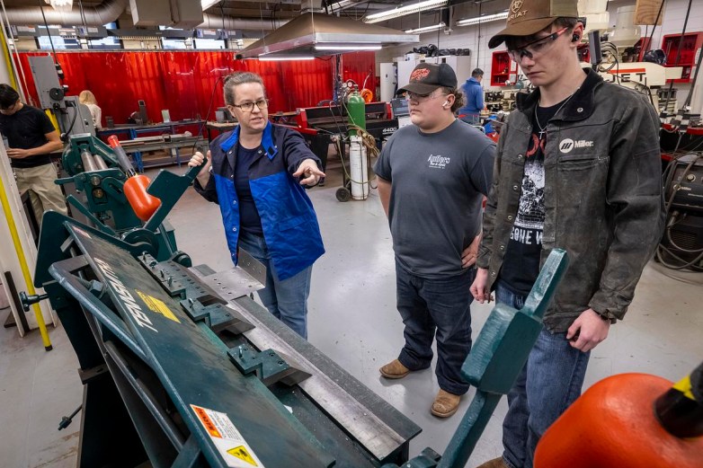 A person wearing a blue jacket gestures and holds a handle of a large metal machine while two other people wearing safety glasses stand beside it in a workshop with tools, tanks and equipment in the background.