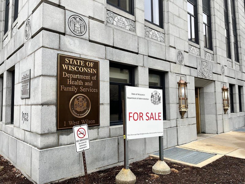 Exterior of a stone building with a sign reading "State of Wisconsin Department of Health and Family Services" and a separate sign reading "FOR SALE" near an entrance.