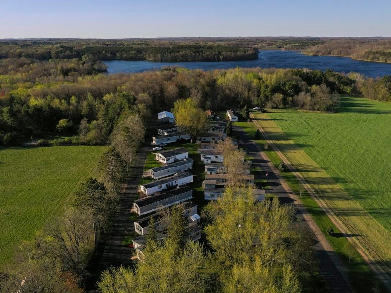 Rows of homes along a road surrounded by trees and open fields, with a lake and forested area in the background.