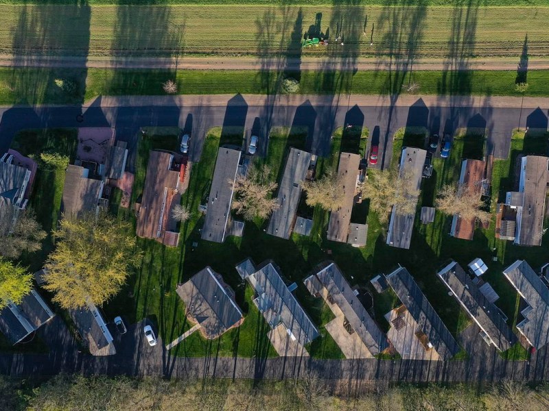 Aerial view of a row of manufactured homes with driveways and cars parked along narrow streets bordered by grass and trees casting long shadows