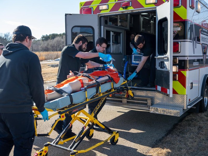 Young men prepare to load a stretcher with a dummy into an ambulance.