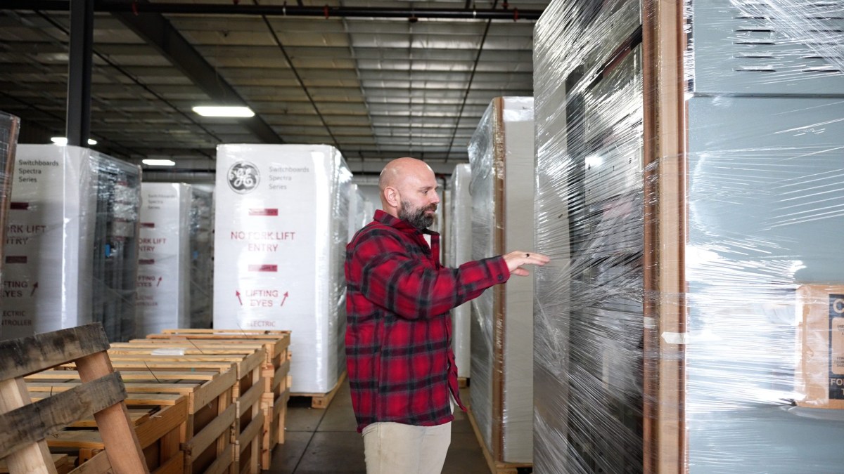 A person in a red plaid shirt stands in a warehouse aisle, extending an arm and hand toward plastic wrap around large boxed equipment, with stacked pallets behind the person.