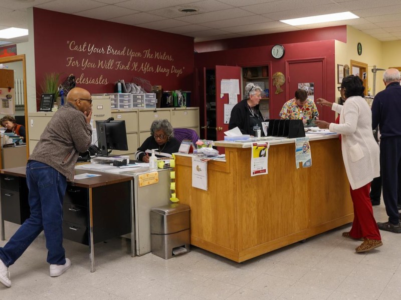 People stand and sit at a front desk area with computers, papers and storage cabinets, with wall text and posters visible in the background.
