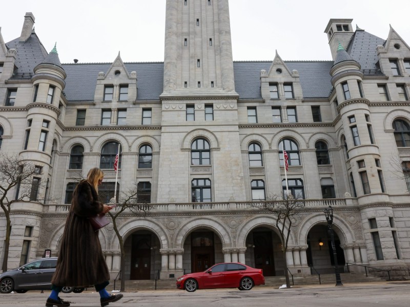 A person walks past a large stone building with arched windows and American flags, looking down at a phone while cars are parked along the street.