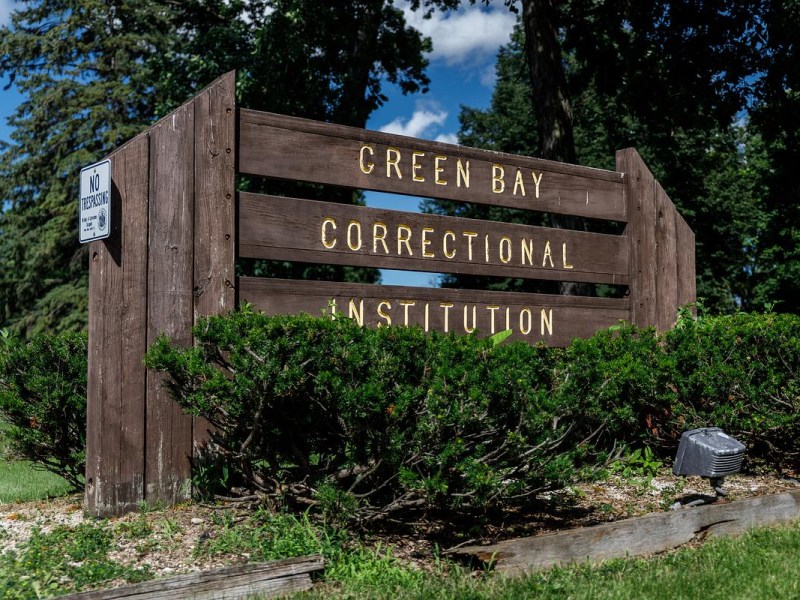 Wooden sign with yellow lettering reads "Green Bay Correctional Institution" beside a smaller "No trespassing" sign, surrounded by green shrubs and trees under a blue sky.