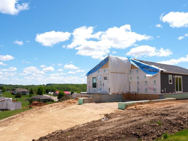 A partially constructed house with exposed insulation and plastic sheeting sits on a dirt lot overlooking a neighborhood under a blue sky with scattered clouds.