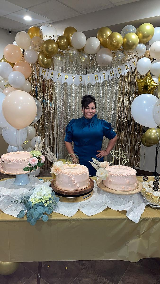 A person stands behind a table with three pink decorated cakes, surrounded by balloons, floral arrangements and a banner reading "HAPPY BIRTHDAY"