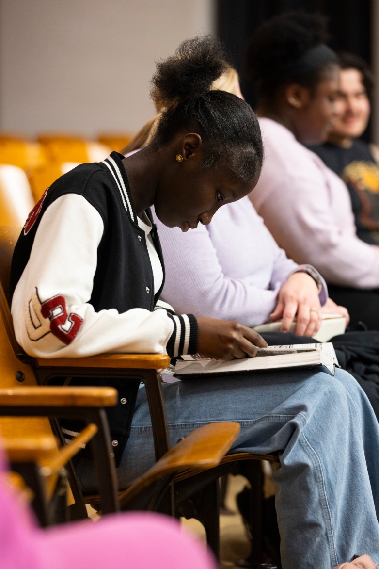 A person sits in a wooden auditorium seat writing in a notebook while others sit in a row beside them holding papers and books