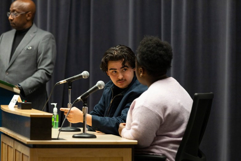Two people sit at a desk with microphones facing each other while a person at a podium stands nearby; a bottle of hand sanitizer sits on the desk beside the microphones