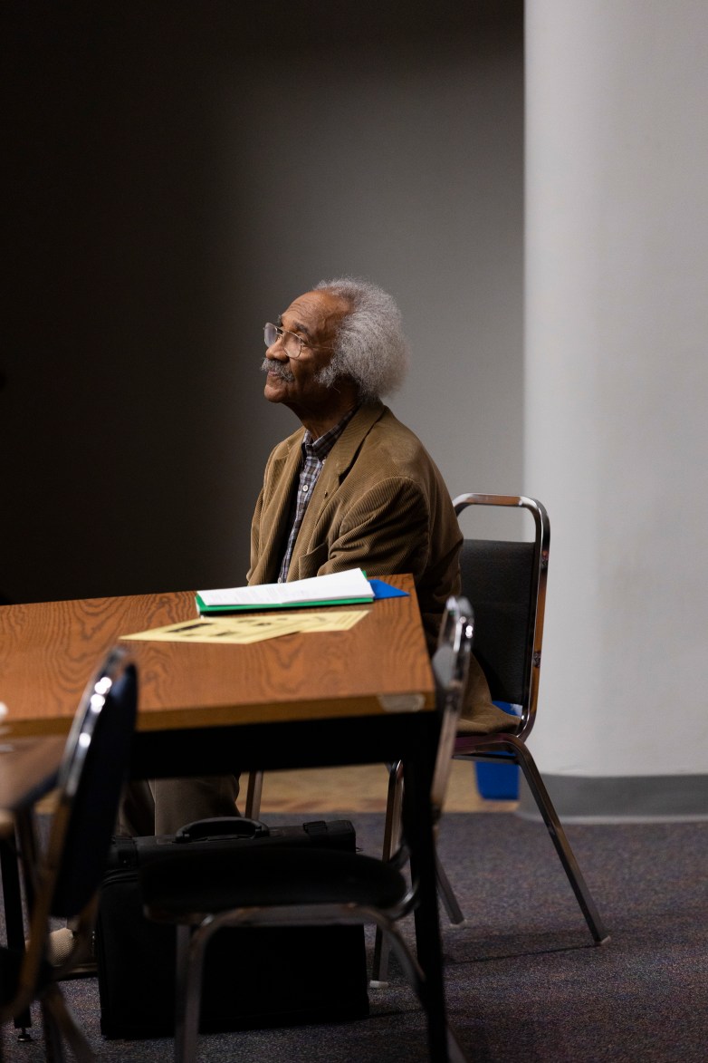 A person with white hair and glasses sits at a table with papers and folders, looking upward; a briefcase rests on the floor beside the chair