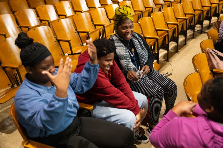 Four people sit in wooden auditorium seats; two raise their hands toward each other while another person in a yellow headwrap holds eyeglasses and a drink cup nearby