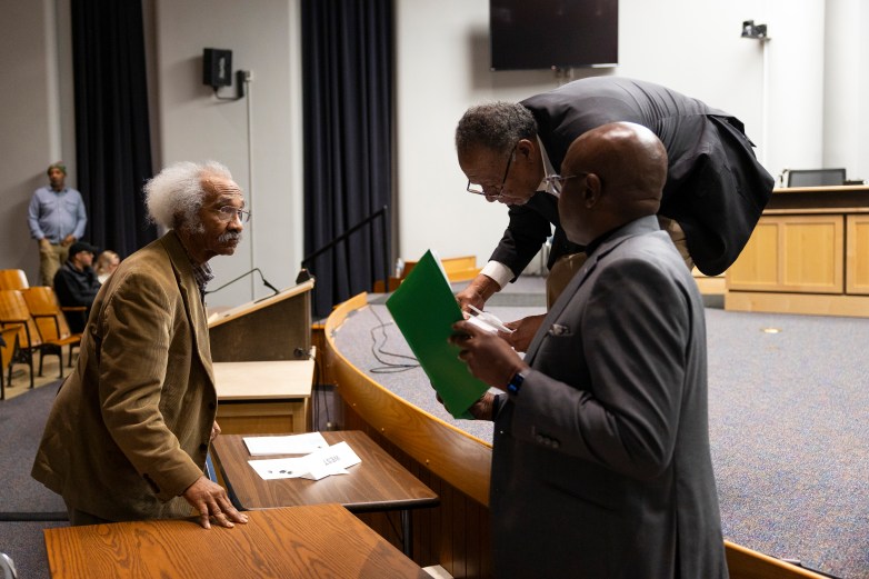 Three people stand and lean over a table in an auditorium; one holds a green folder while another rests a hand on the table near scattered papers