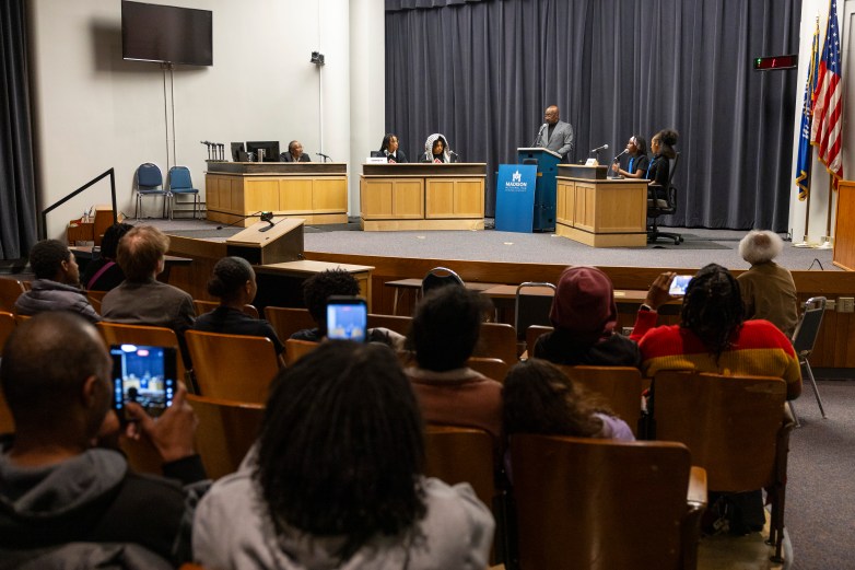 An audience sits facing a stage where several people sit behind desks with microphones while another person stands at a podium labeled "Madison Metropolitan School District"