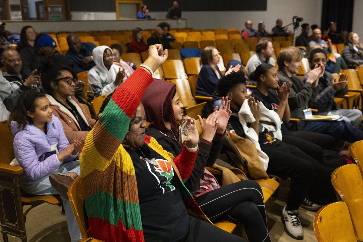 People sit in wooden auditorium seats clapping while one person in the foreground raises a fist and holds a phone, with others seated in rows behind them