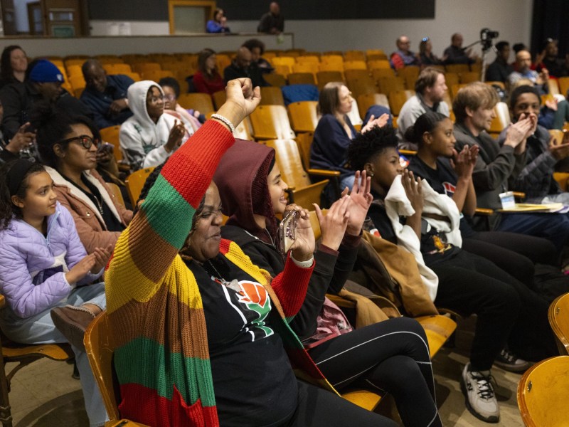 People sit in wooden auditorium seats clapping while one person in the foreground raises a fist and holds a phone, with others seated in rows behind them