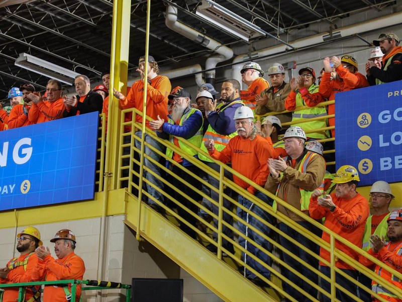 Workers in hard hats and yellow and orange vests clap inside a building