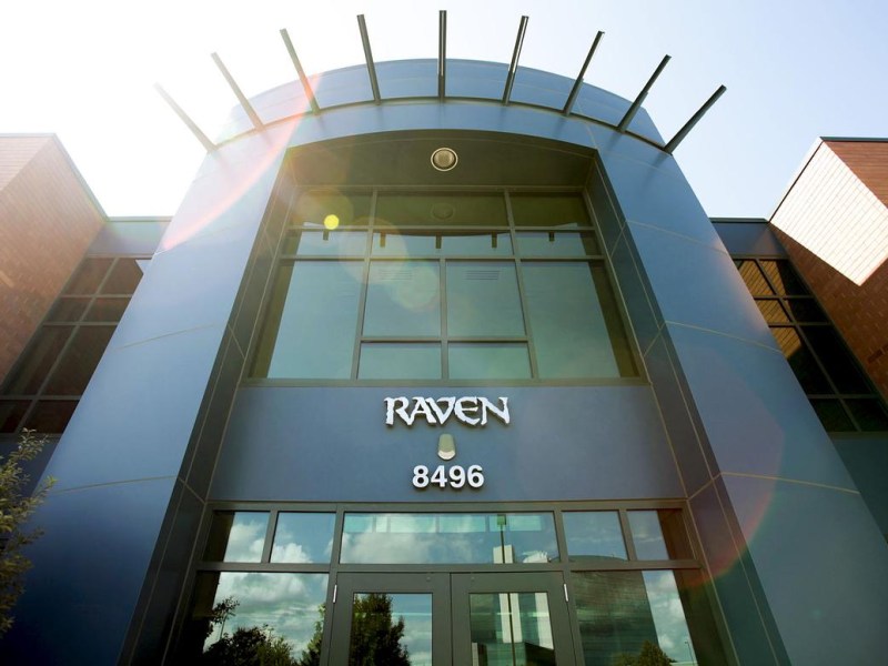 A building entrance labeled "Raven" is show from a low angle, stretching into the sky. The entrance is mostly windows, with blue trim. Rays of sunshine are shown on the upper lefthand of the building.