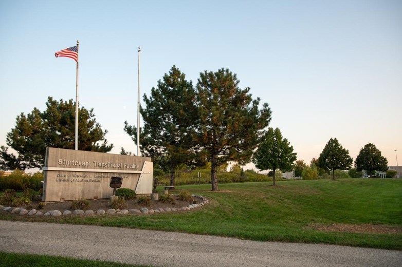 A concrete sign reading "Sturtevant Transitional Facility" stands beside two flagpoles and a row of trees along a grassy area.