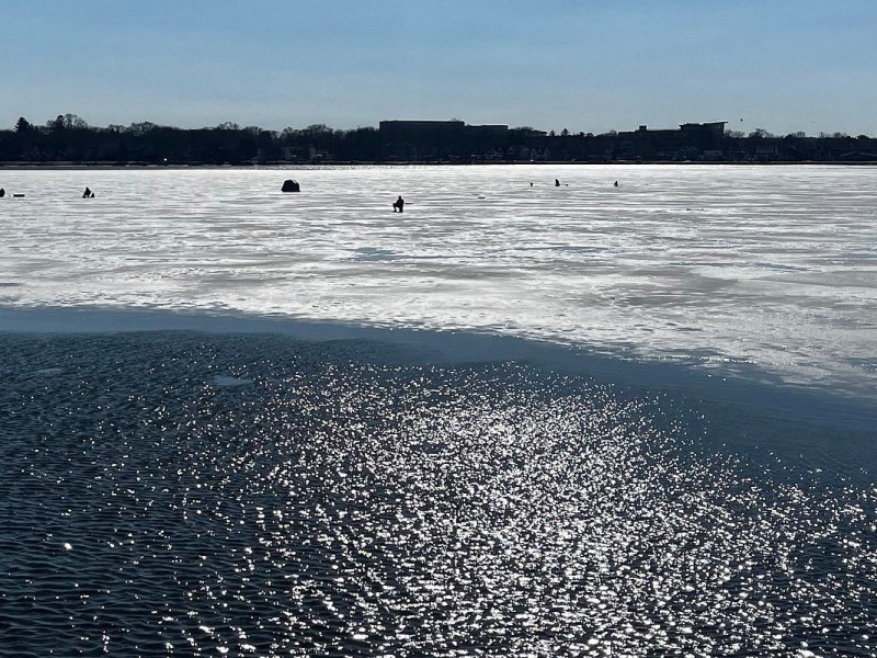 Open water ripples in the foreground as people and small shelters sit scattered across a snow-covered frozen lake, with buildings and trees along the far shoreline.