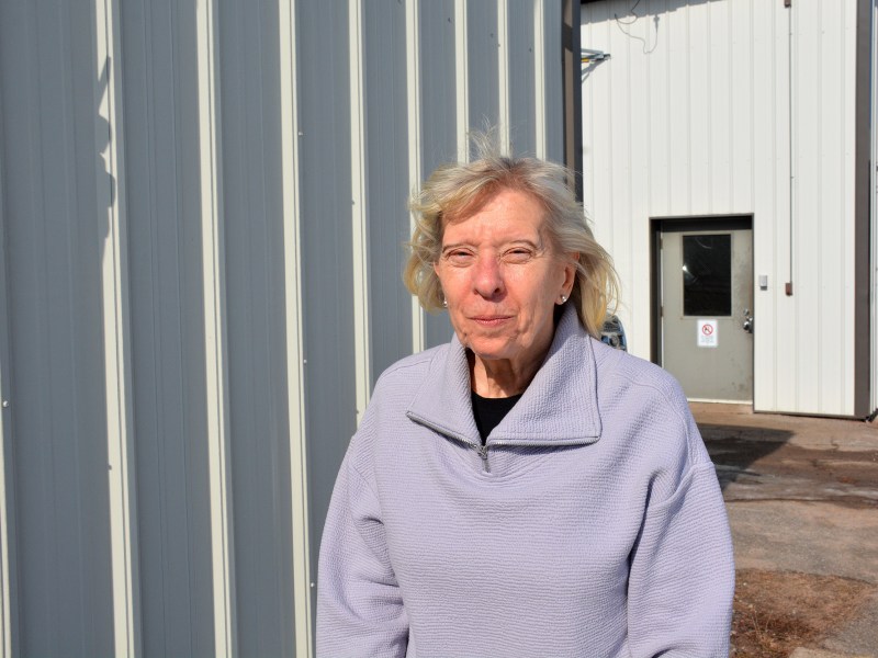 A person wearing a lavender pullover stands outside near a metal-sided building, with a closed door and concrete walkway visible behind the person.