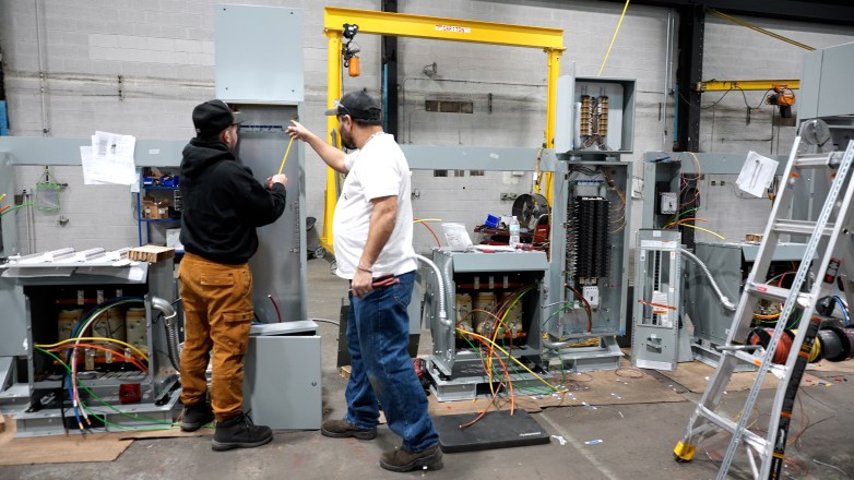 Two people stand in a workshop beside open electrical cabinets and wiring, with one person holding a tape measure, and tools and a ladder are nearby.