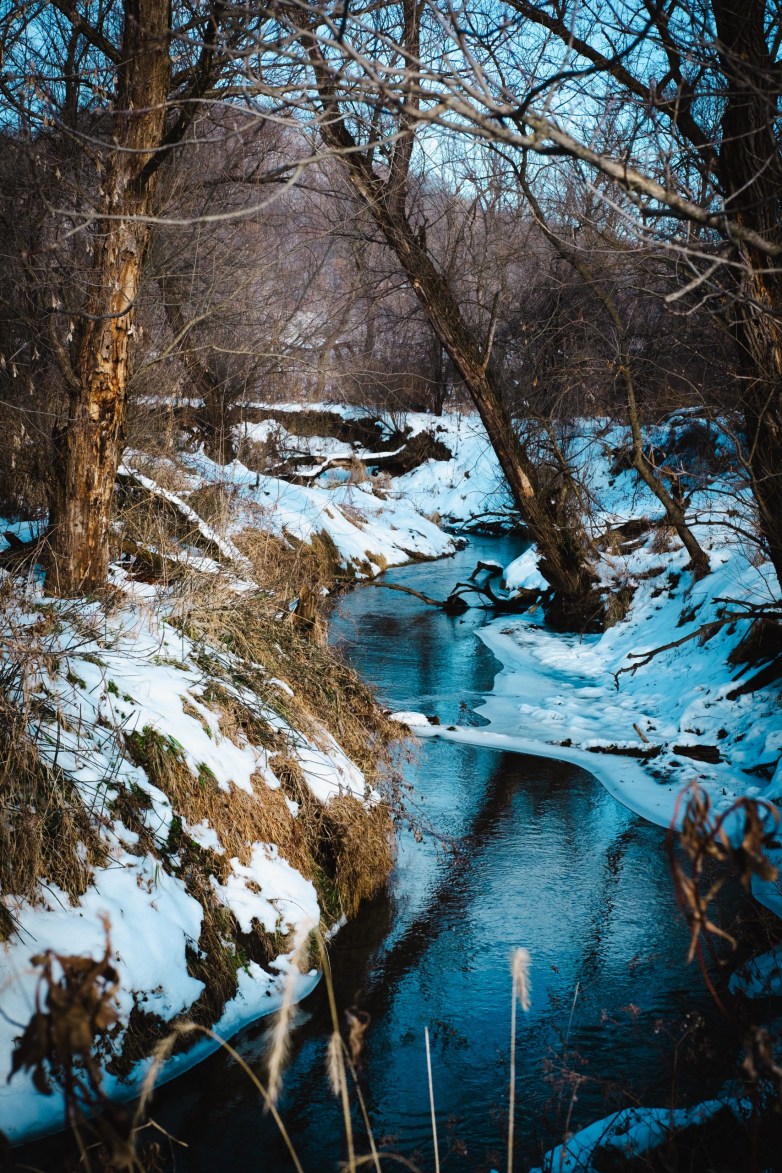 A narrow creek winds between snow-covered banks and leafless trees, with patches of ice along the water in a wooded area.