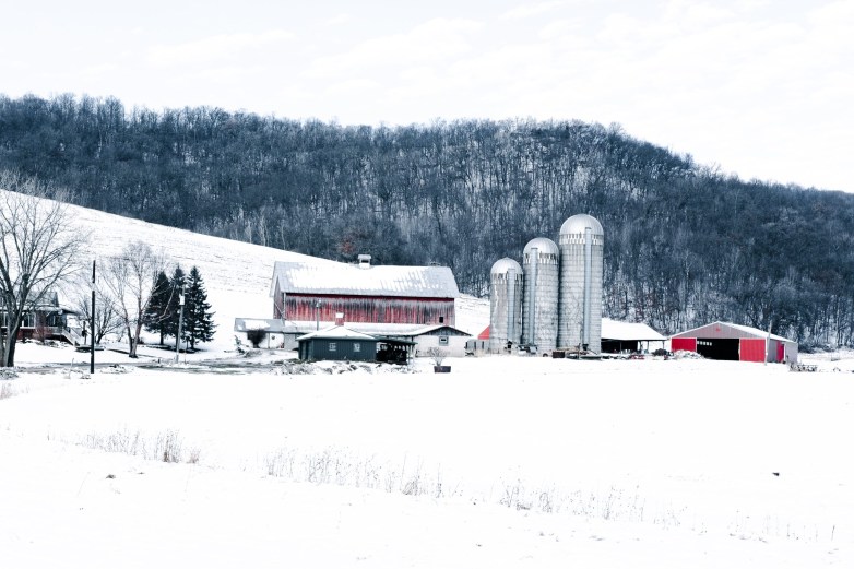 A farm sits in a snow-covered valley with a red barn, three tall silos and outbuildings near a wooded hillside.